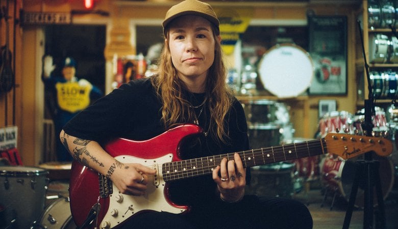 Rebecka Mardal sitter på en stol och spelar på en rödvit gitarr i en lokal. Hon tittar in i kameran och har en keps på sig.