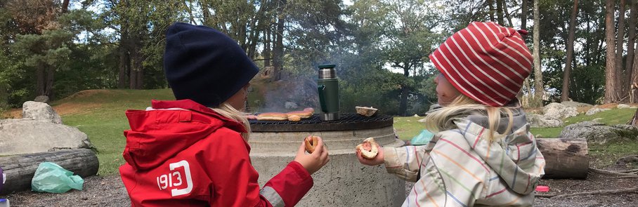 Några barn sitter på toppen av en stock och äter korv vid en grillplats i skogen.