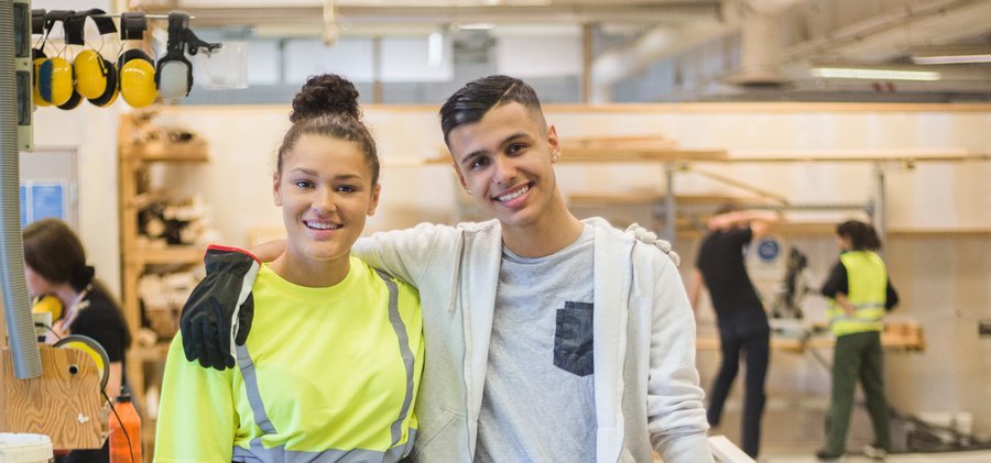 Full length portrait of smiling female trainee standing with arm around male coworker at workbench