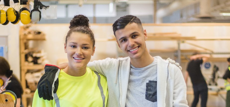 Full length portrait of smiling female trainee standing with arm around male coworker at workbench