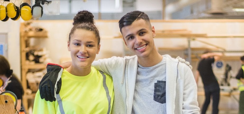 Full length portrait of smiling female trainee standing with arm around male coworker at workbench