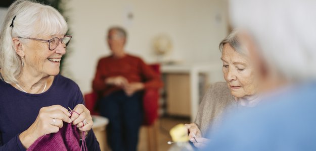 Senior female friends spending leisure time knitting while sitting at table in retirement home