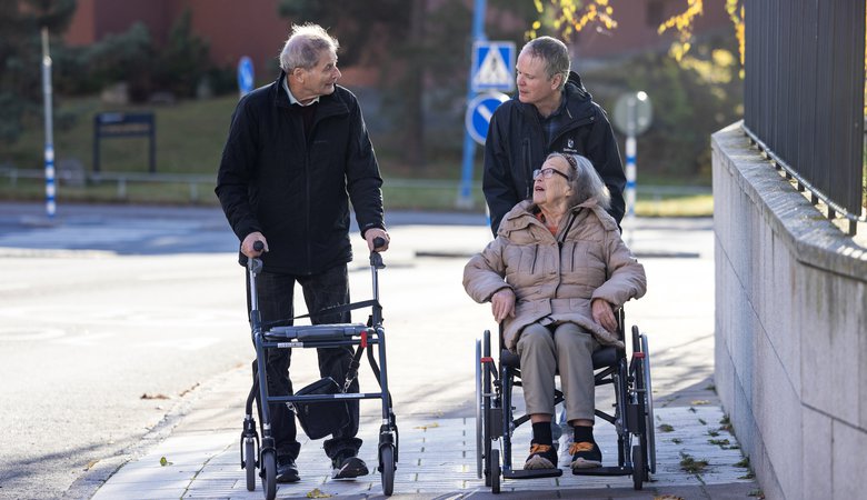 Äldre man med rullator och äldre kvinna i rullstol på promenad i Edsberg tillsammans med vårdbiträde.